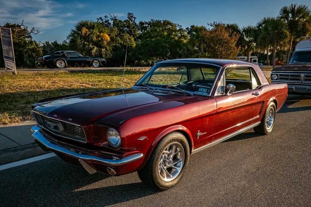 A red 1965 Red Ford Mustang. Its parked on a road next to a grassfield